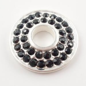 Könnte beinhalten: Eine silberfarbene, kreisförmige Perle mit einem zentralen Loch. Die Perle ist mit Reihen kleiner, schwarzer, facettierter Steine besetzt. Das Design ist symmetrisch, wobei die schwarzen Steine eine glitzernde, strukturierte Oberfläche bilden.