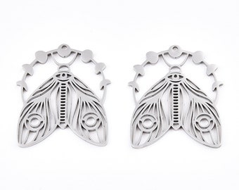 May include: Two silver-colored moth-shaped pendants. Each pendant features intricate wing detailing, a central body with vertical lines, and circular eye-like designs. A decorative arc with small circles and curved elements surrounds the moth's head.