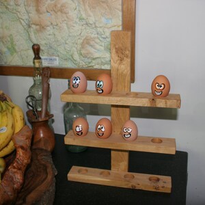 May include: A wooden egg holder with six brown eggs, each with a different hand-painted face. The holder has two shelves and is made of natural wood.