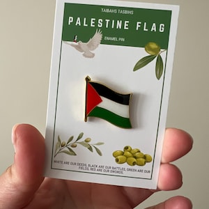 Puede incluir: Un pin de esmalte con la bandera de Palestina, con colores rojo, negro, blanco y verde. El pin está en una tarjeta blanca con el texto "PALESTINE FLAG" y "ENAMEL PIN". La tarjeta también tiene ramas de olivo y aceitunas.
