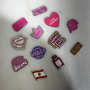 May include: A selection of enamel pins in diverse shapes and colours. The pins feature phrases such as "XOXO", "Love Yourself", and "Good Vibes Only". Other pins depict a ticket, a crown, a baseball cap, and a perfume bottle.