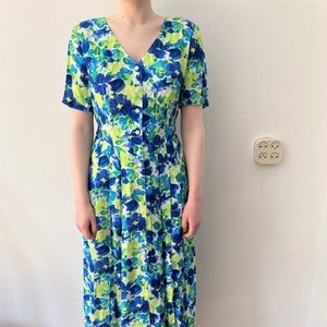 Op de afbeelding: Een witte midi-jurk met een blauwe en groene bloemenprint. De jurk heeft een v-hals en korte mouwen. Het heeft een knoopsluiting aan de voorkant en een geplooide rok.