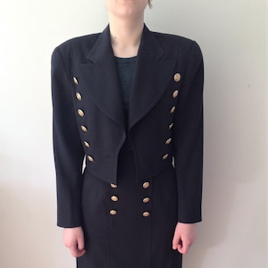 Op de afbeelding: Een zwart tweedelig pak met een korte jas en een knielange rok. De jas heeft een dubbelknoops ontwerp met goudkleurige knopen. De rok heeft ook gouden knopen. Het pak is een klassieke, getailleerde stijl.