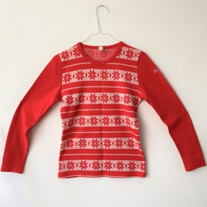 May include: A red long-sleeved shirt with a white snowflake pattern across the chest. The shirt is hanging on a wire hanger against a white background. The sleeves and back are solid red.