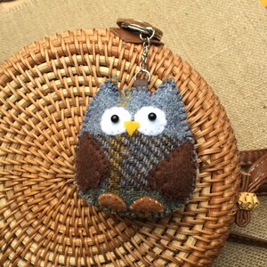 May include: A blue and brown owl keychain made of felt with a plaid pattern. The owl has white eyes and a yellow beak. The keychain is attached to a silver chain and a brown leather loop.