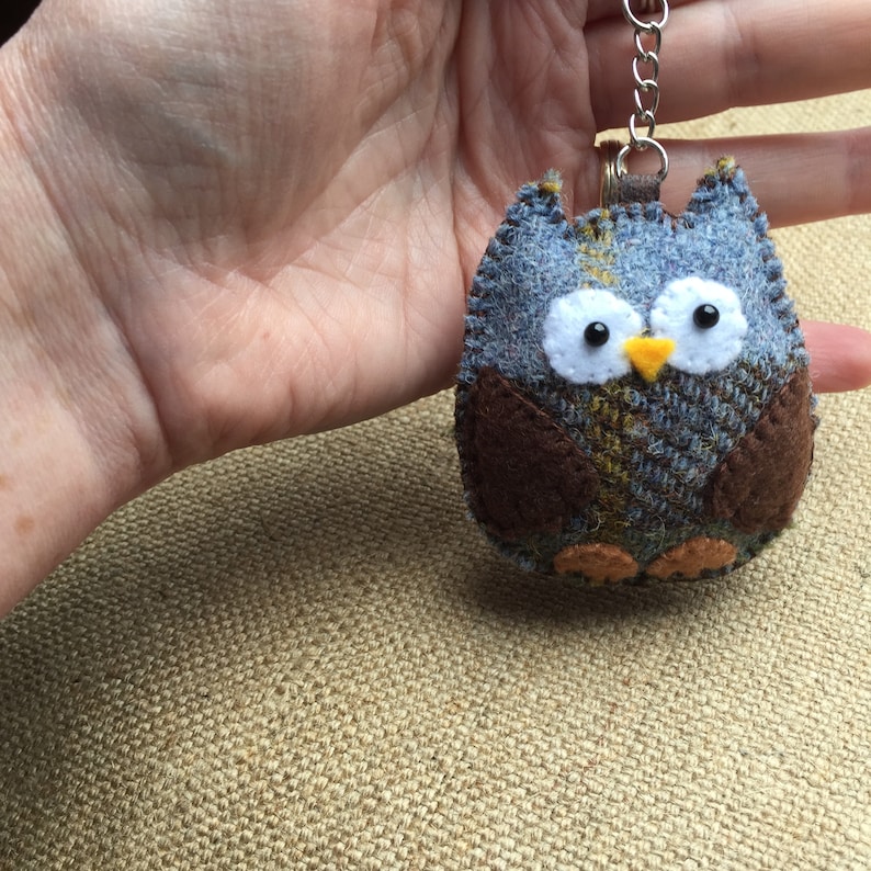 May include: A blue and brown owl keychain made of tweed fabric. The owl has white eyes with black pupils and a yellow beak.