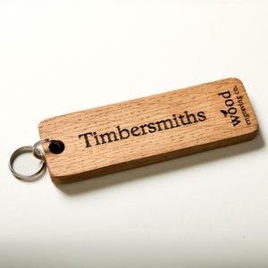 May include: A rectangular light brown wooden keyring featuring a metal ring attached through a circular hole. The wood surface is engraved with the text Timbersmiths and the logo wood engraving co. in a dark brown font with visible grain.