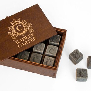 May include: A wooden box with a personalized engraved lid that says "Radley Carter" with a crest and the letter "C". The box contains nine gray stone cubes with the letter "C" engraved on each. Three additional cubes are outside the box.