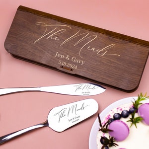 May include: A wooden box with a personalized inscription "The Meads Jen & Gary 5.18.2024" and two silver cake servers with the same inscription. The servers are on a pink surface with a cake in the background.