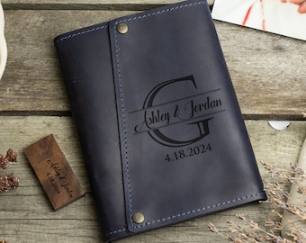 May include: A dark blue leather journal with a personalized monogram and the names "Ashley & Jordan" and the date "4.18.2024" engraved on the front.