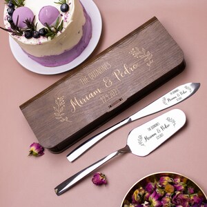 Op de afbeelding: Een houten doos met een gouden gegraveerd ontwerp met de namen "Miriam & Pedro" en de datum "12.9.2023". De doos bevat een zilveren taartmes en een serverset met bijpassende gegraveerde ontwerpen.