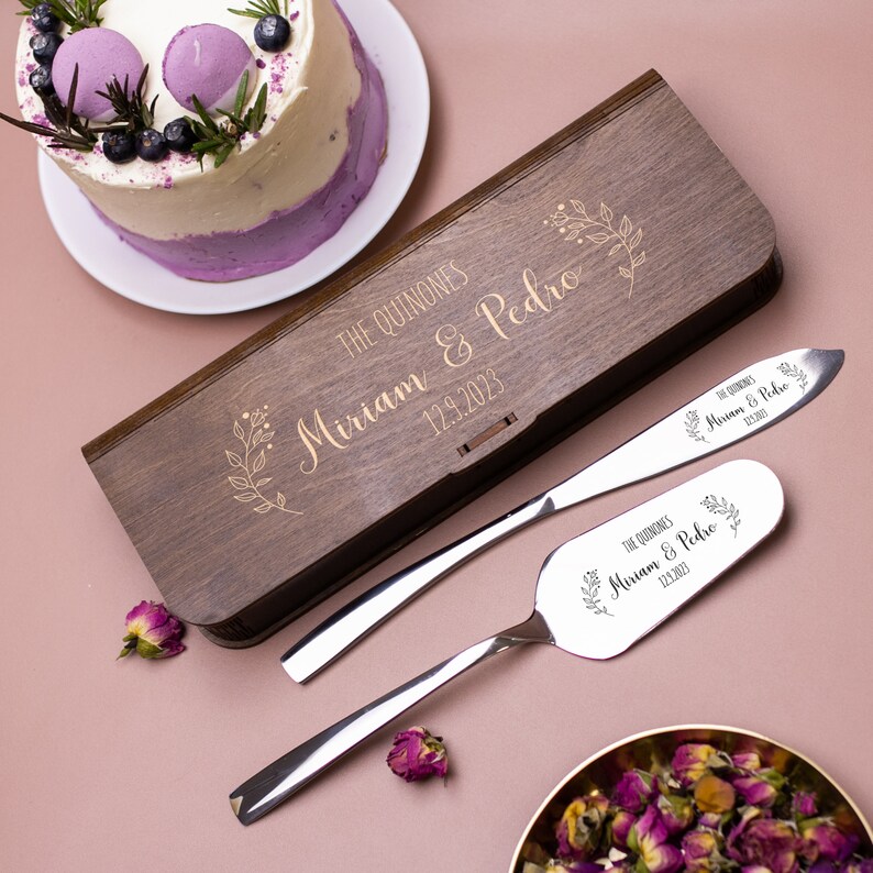 Op de afbeelding: Een houten doos met een gouden gegraveerd ontwerp met de namen "Miriam & Pedro" en de datum "12.9.2023". De doos bevat een zilveren taartmes en een serverset met bijpassende gegraveerde ontwerpen.