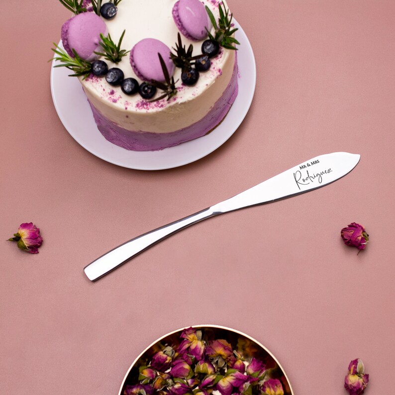 May include: A silver cake knife with the inscription "Mr. & Mrs. Rodriguez" on a pink background with a purple and white cake and pink rose buds.