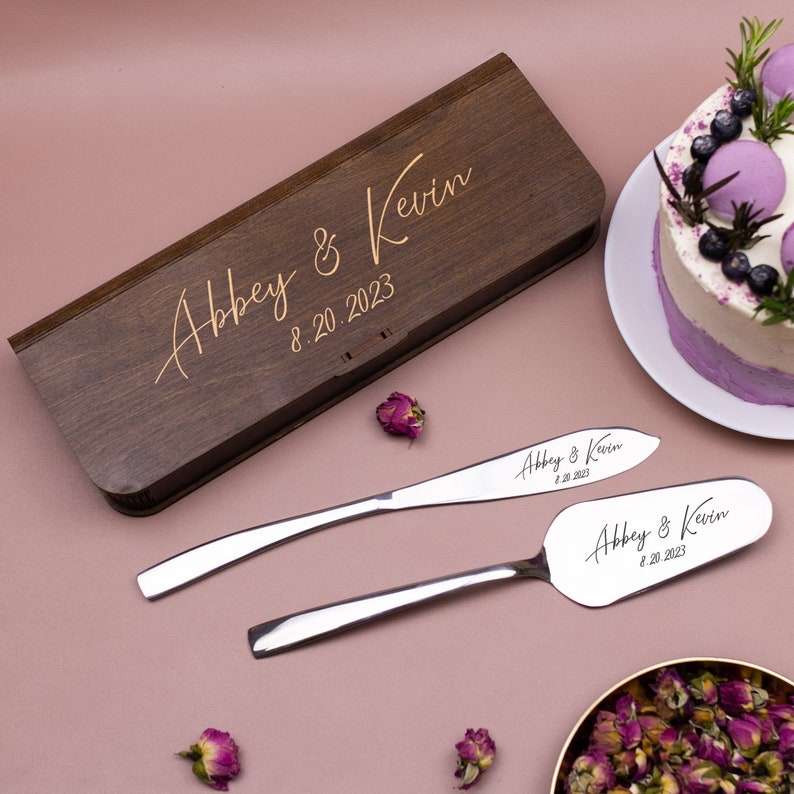 May include: A wooden box with the inscription "Abbey & Kevin 8.20.2023" and two silver cake knives with the same inscription.