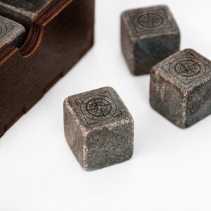 May include: Four gray stone cubes with a compass design etched on each side. The cubes are sitting on a white surface next to a wooden box.