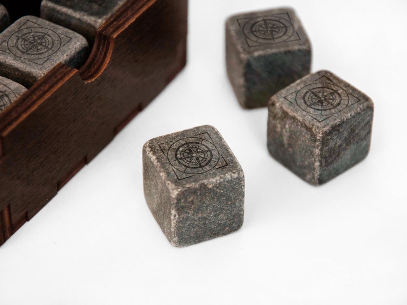 May include: Four gray stone cubes with a compass design etched on each side. The cubes are sitting on a white surface next to a wooden box.