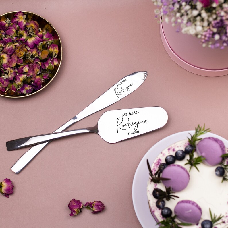 May include: A silver cake server and knife set with the text "MR. & MRS. Rodriguez 11.22.2023" engraved on the handles. The set is on a pink surface with a cake and flowers in the background.