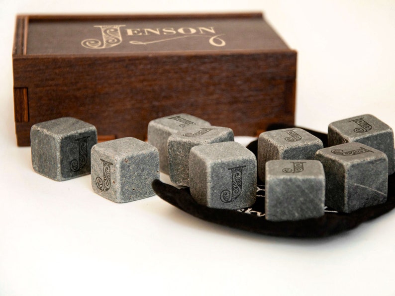May include: A set of gray stone whiskey chilling cubes with the letter "J" engraved on each cube. The cubes are in a black velvet pouch and come in a wooden box with the name "Jenson" engraved on the top.