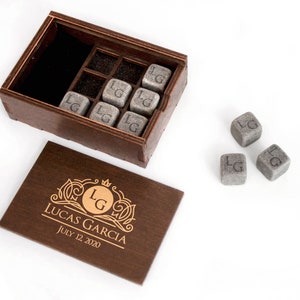 May include: A wooden box with a lid that has a personalized inscription. The inscription reads "Lucas Garcia July 12, 2020". The box contains eight gray stone cubes with the initials "LG" engraved on each cube. Three additional cubes are outside the box.