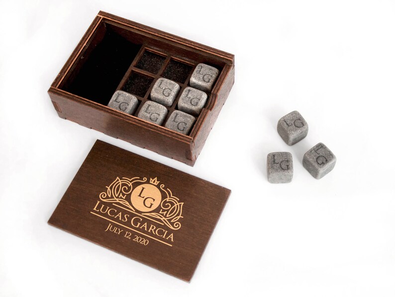 May include: A wooden box with a lid that has a personalized inscription. The inscription reads "Lucas Garcia July 12, 2020". The box contains eight gray stone cubes with the initials "LG" engraved on each cube. Three additional cubes are outside the box.