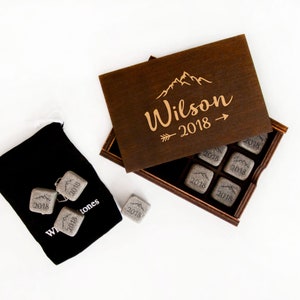 May include: A wooden box with a lid engraved with the text "Wilson 2018" and a mountain range design. The box contains eight gray stone cubes, each engraved with the year "2018".