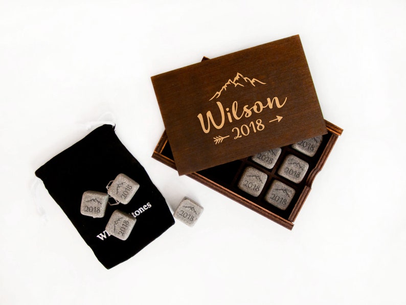 May include: A wooden box with a lid engraved with the text "Wilson 2018" and a mountain range design. The box contains eight gray stone cubes, each engraved with the year "2018".