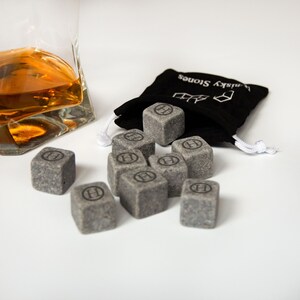 May include: A set of gray whiskey stones in a black bag with the text "Whiskey Stones" printed on it. The stones are shaped like cubes and have a letter "H" engraved on each side.