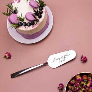 May include: A silver cake server with the inscription "Abbey & Kevin 8.20.2023" on a pink background.