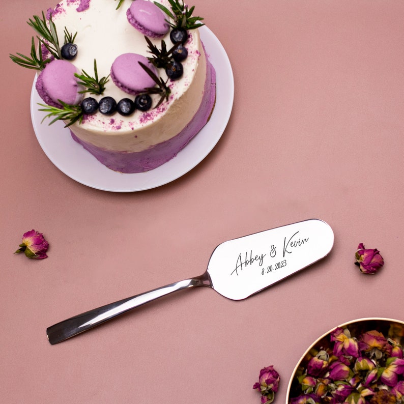 May include: A silver cake server with the inscription "Abbey & Kevin 8.20.2023" on a pink background.