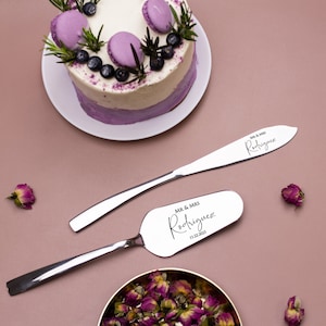 May include: A silver cake knife and server set with the text "MR & MRS Rodriguez 11.22.2023" engraved on the handles. The set is on a pink background with a purple and white cake and a small bowl of dried rose petals.