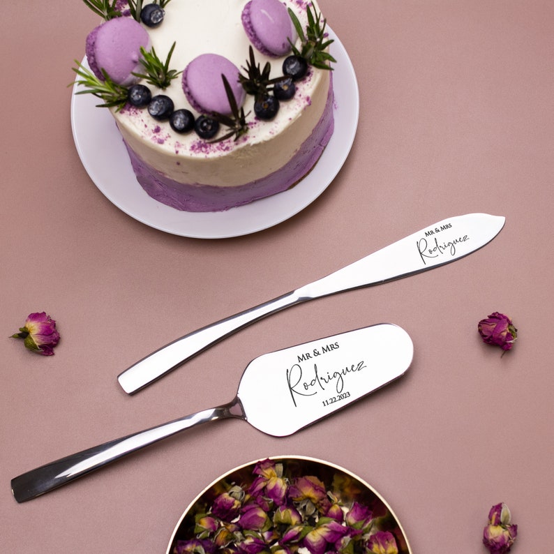 May include: A silver cake knife and server set with the text "MR & MRS Rodriguez 11.22.2023" engraved on the handles. The set is on a pink background with a purple and white cake and a small bowl of dried rose petals.
