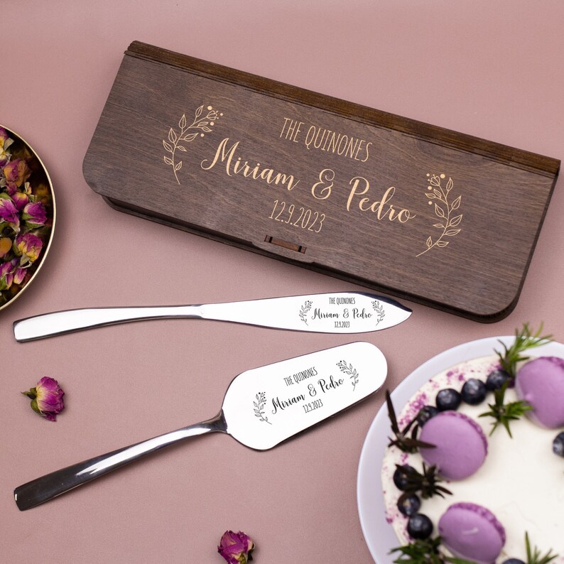 Op de afbeelding: Een houten doos met een gouden gegraveerd ontwerp met de namen Miriam & Pedro en de datum 12.9.2023. De doos bevat twee zilveren taartspaten met hetzelfde gegraveerde ontwerp.