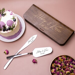 May include: A wooden box engraved with "Abbey & Kevin 8.20.2023" and two silver cake servers with the same inscription. The servers are on a pink surface with a purple and white cake in the background.