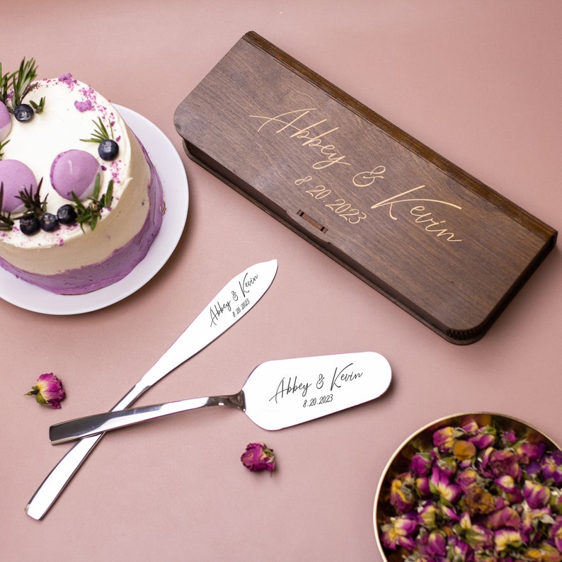 May include: A wooden box engraved with "Abbey & Kevin 8.20.2023" and two silver cake servers with the same inscription. The servers are on a pink surface with a purple and white cake in the background.