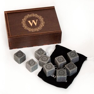 May include: A set of eight gray whiskey stones in a black velvet bag with a wooden box. The stones are engraved with a wreath and the letter "W".