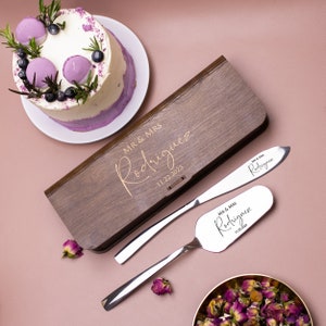 May include: A wooden box with a gold inscription "MR & MRS Rodriguez 11.22.2023" and two silver cake knives with the same inscription. The knives are in a wooden box.