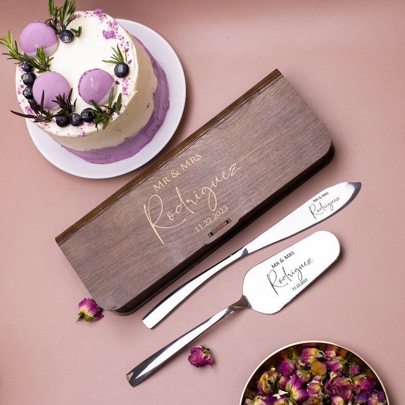 May include: A wooden box with a gold inscription "MR & MRS Rodriguez 11.22.2023" and two silver cake knives with the same inscription. The knives are in a wooden box.