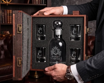 May include: A dark wooden presentation box containing a clear glass decanter and four matching glasses. The decanter and glasses are etched with a crown emblem and the letter C. The decanter also features the text RADLEY CARTER.