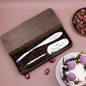Op de afbeelding: Een houten doos met een bruine afwerking bevat twee zilveren taartspaten met gegraveerde bloemmotieven en de tekst "The Quinn's Miriam & Pedro 12.18.2023".