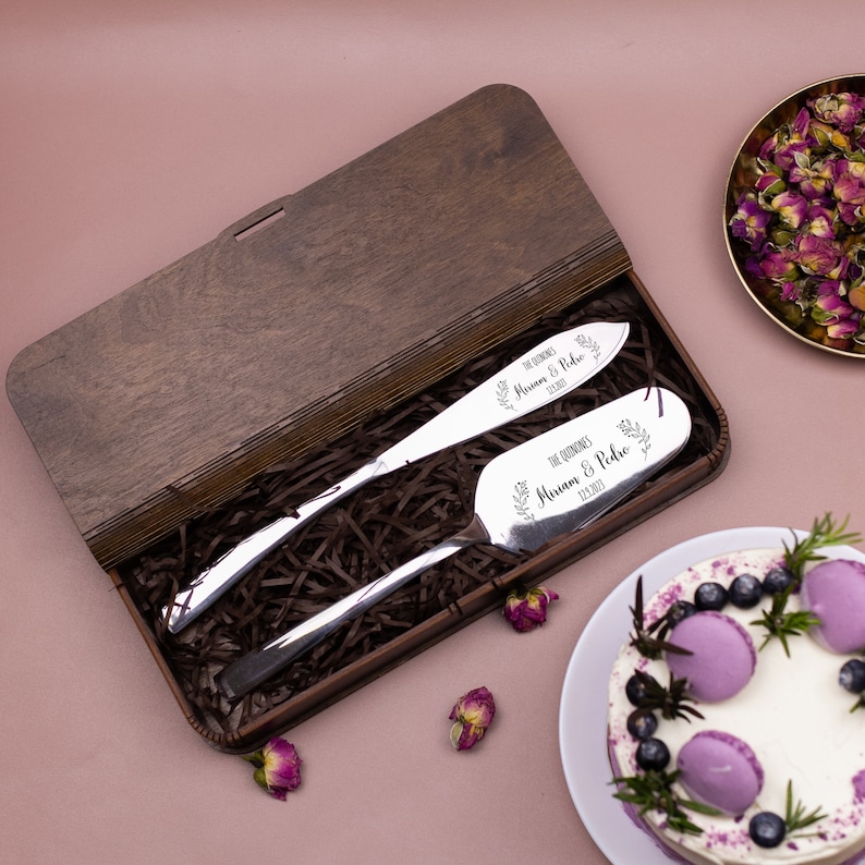 Op de afbeelding: Een houten doos met een bruine afwerking bevat twee zilveren taartspaten met gegraveerde bloemmotieven en de tekst "The Quinn's Miriam & Pedro 12.18.2023".