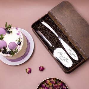 May include: A wooden box with a brown finish contains two silver cake servers. The servers are engraved with the names "Abbey & Kevin" and the date "8.28.2023".