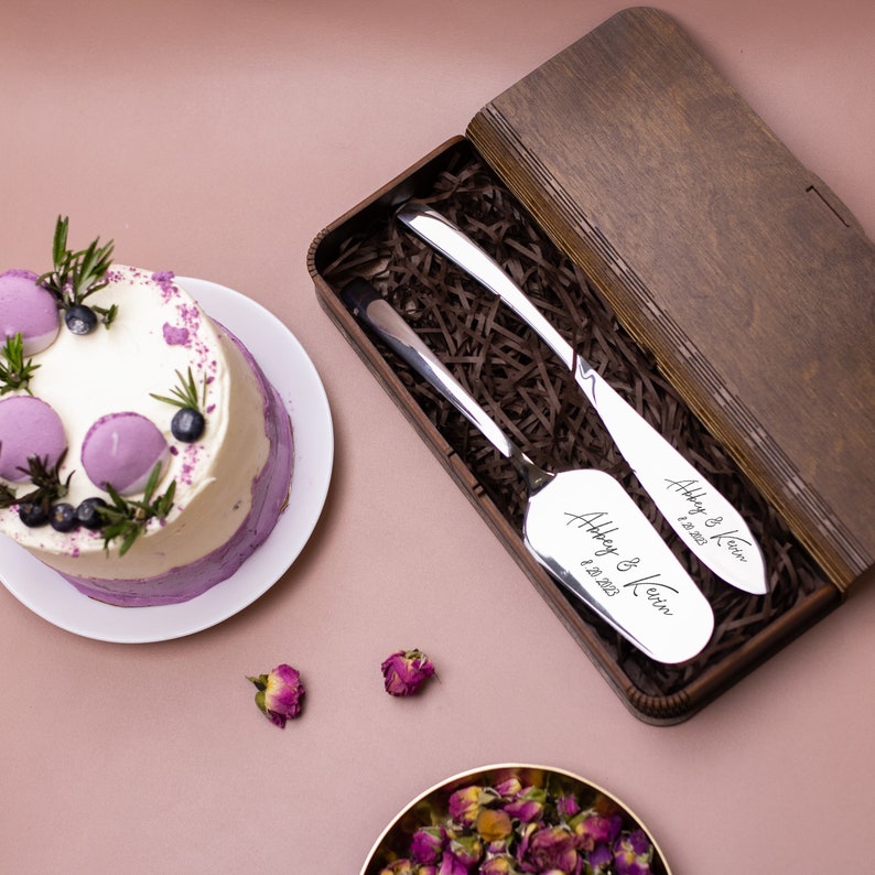 May include: A wooden box with a brown finish contains two silver cake servers. The servers are engraved with the names "Abbey & Kevin" and the date "8.28.2023".