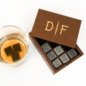 May include: A wooden box with the initials "D.F." engraved on the lid. The box contains nine gray stone cubes, each engraved with the initials "D.F.". Three of the cubes are in a glass with amber liquid.