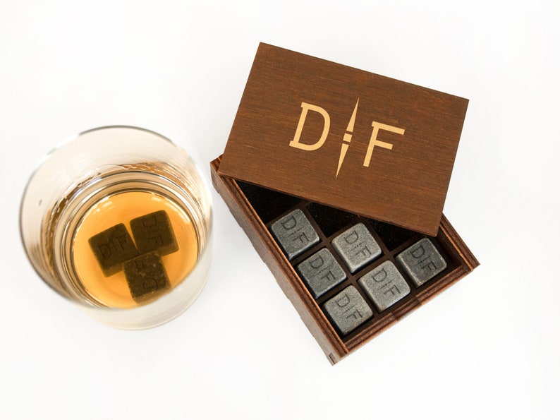 May include: A wooden box with the initials "D.F." engraved on the lid. The box contains nine gray stone cubes, each engraved with the initials "D.F.". Three of the cubes are in a glass with amber liquid.