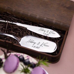 May include: A wooden box with brown paper shredding inside. Two silver cake servers are in the box. The servers are engraved with the names "Abbey & Kevin" and the date "8.20.2023".
