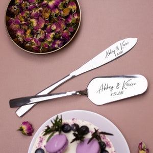 May include: A silver cake knife and server set with the text "Abbey & Kevin 8.20.2023" engraved on each piece. The set is on a pink surface with a bowl of dried rose petals in the background.