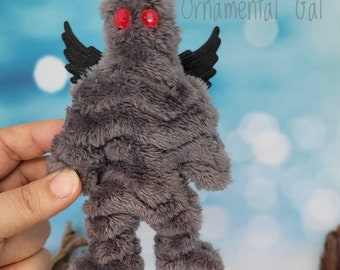 May include: Gray plush toy with black wings and red eyes. The toy is standing on a surface with a blue background.