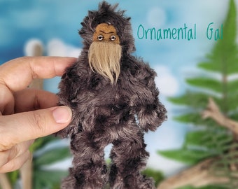May include: A brown plush toy resembling a Bigfoot with a long, blonde beard. The toy has a human-like face with brown eyes and a furrowed brow. The toy is being held in a hand against a blurred background of green foliage and a blue sky.