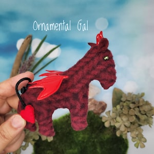 May include: A small, red plush toy shaped like a horse with a red devil's wing and a heart-shaped tail. The toy has a green gemstone eye and is standing on a green surface.