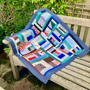 May include: A colorful patchwork quilt with a blue border rests on a wooden bench. The quilt features a variety of fabrics in shades of blue, green, pink, and brown, arranged in a geometric pattern. The quilt is approximately 40 inches square.